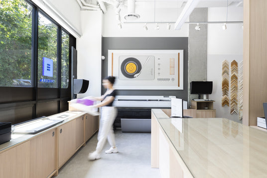 A person in motion carries a framed item inside a bright, modern art shop with large windows, wood accents, and framed artwork on the wall. Framing samples are displayed on the right side.