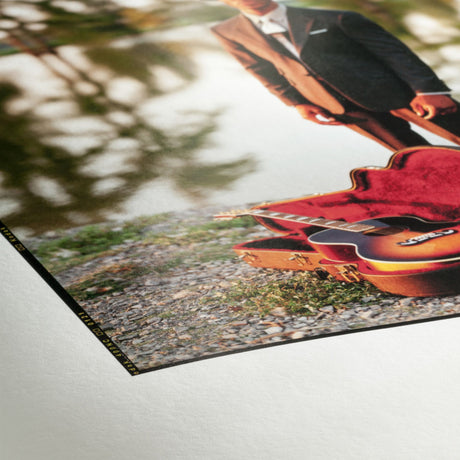 A photograph printed on Hahnemühle FineArt Pearl by Hahnemuhle features a person in a suit outdoors next to an open guitar case and guitar on gravel, with trees reflected in the background.
