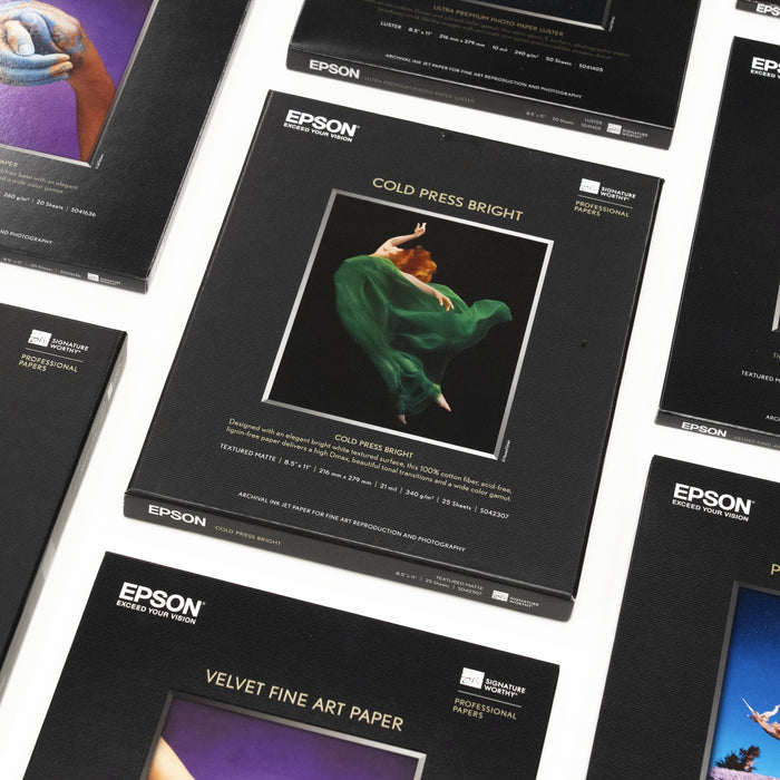Multiple Epson professional paper packages are arranged on a white surface. The central package features Cold Press Bright with an image of a dancer in a green dress mid-air. Other labeled papers are partially visible.