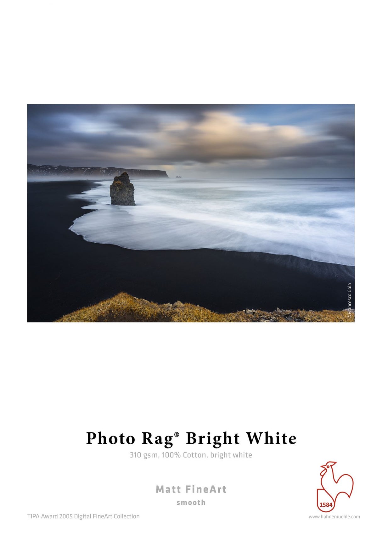 A long-exposure of a rocky sea stack and black sand beach with white waves beneath a cloudy sky, printed on Hahnemuhle Photo Rag® Bright White by Hahnemuhle for exceptional FineArt quality.