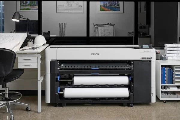 A large Epson wide-format printer with two rolls of paper sits in an office, surrounded by a chair, desk with a hard hat, architectural drawings, and shelves with more rolled paper and supplies.